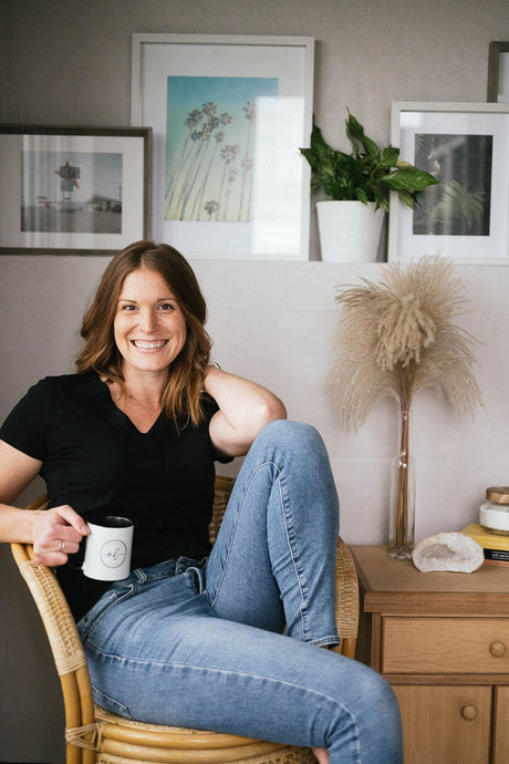 Women sitting on chair wear jeans and a black t-shirt with one elbow rested on bent knee and holding a coffee. 